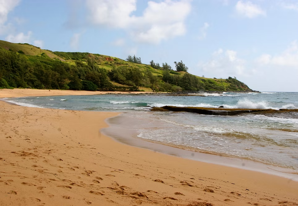 Plage de sable doré avec des vagues et une côte verte avec des arbres et des collines sous un ciel partiellement nuageux.