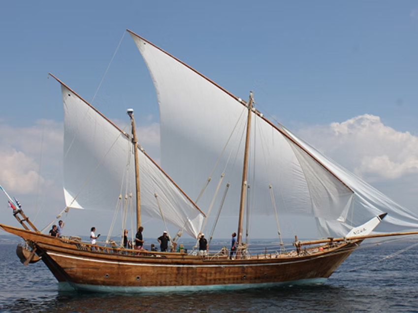 Un voilier en mer avec plusieurs personnes à bord et des voiles déployées contre un ciel partiellement nuageux.