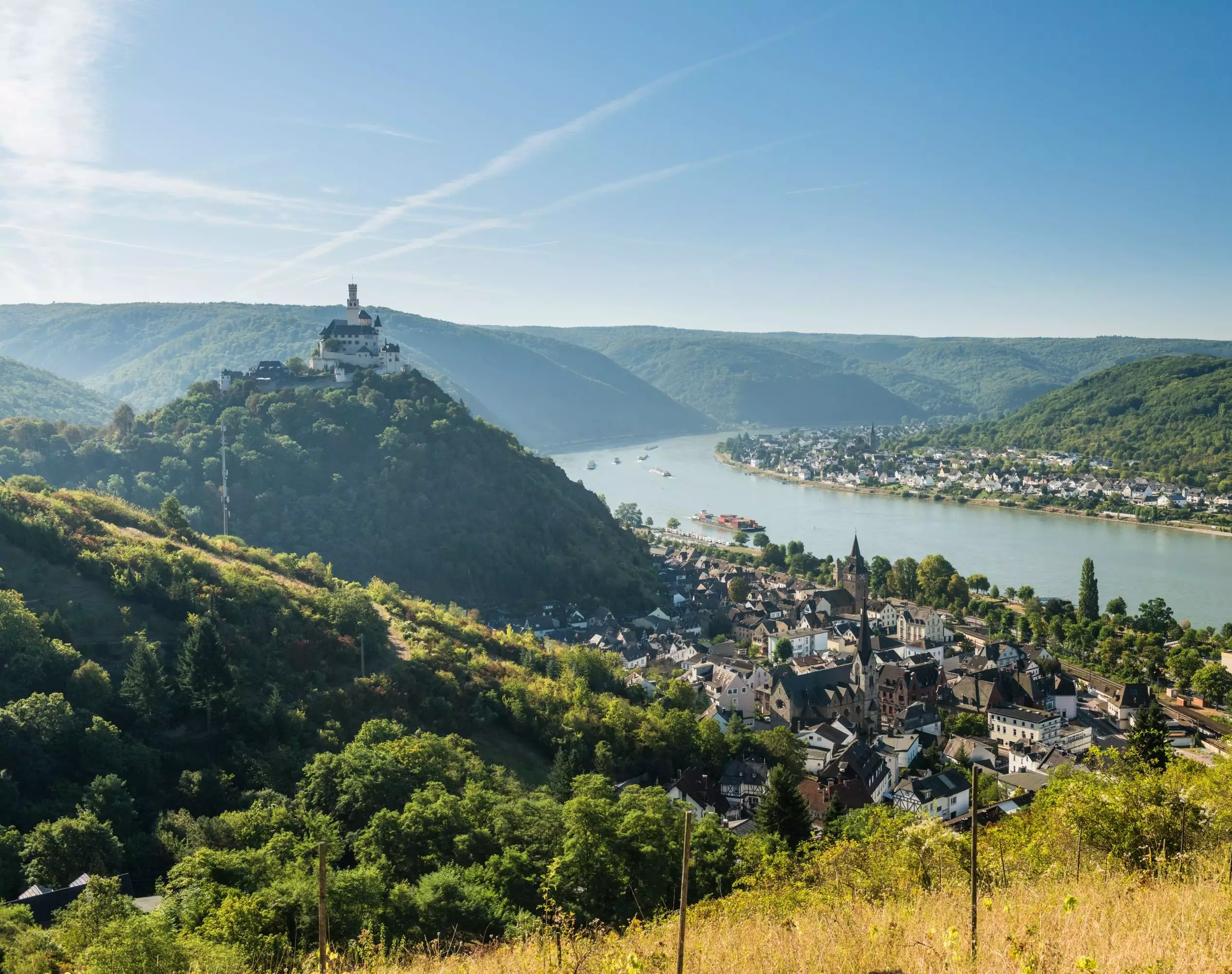 Vue panoramique d'une vallée traversée par une rivière, avec une ville en bord de l'eau et un château sur une colline verdoyante, sous un ciel bleu clair.