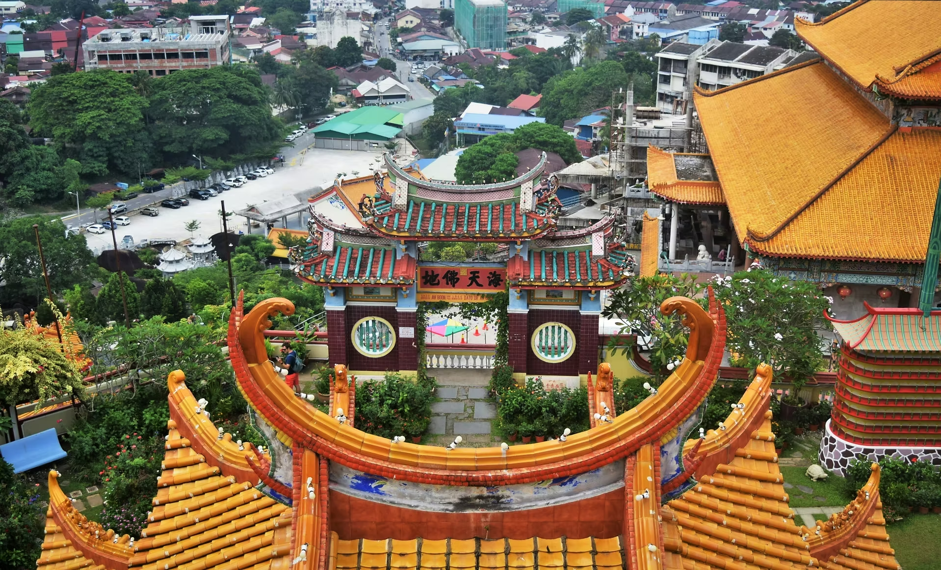 Vue aérienne d'un temple chinois avec un portail orné de dragons, des toits orange et verts, entouré de végétation verte et d'une ville en arrière-plan.