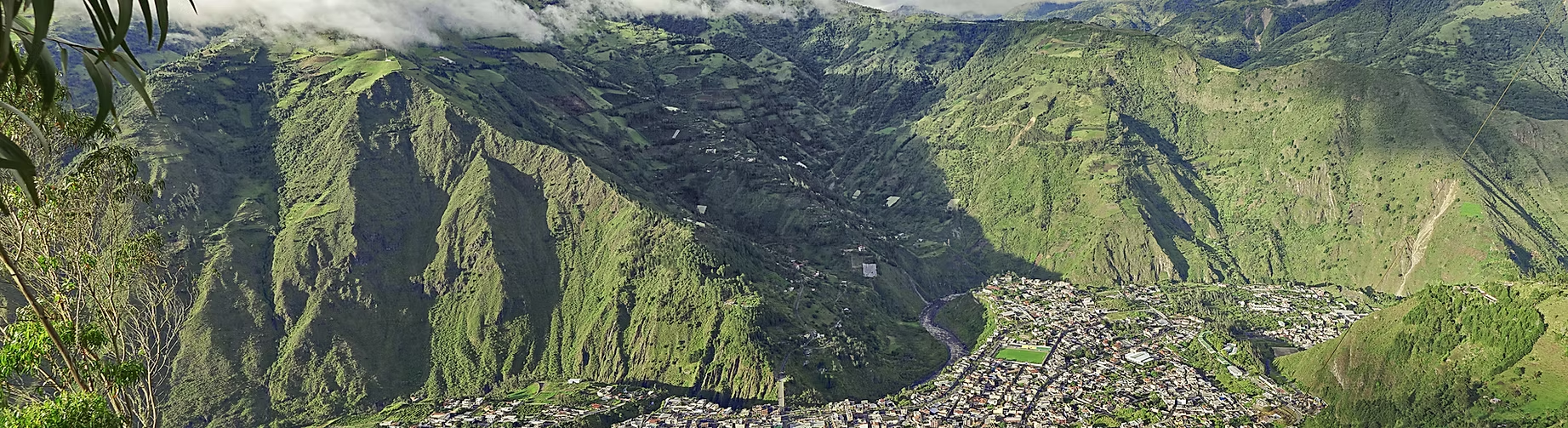 Vue panoramique d'une ville au fond de vallées entourées de montagnes vertes, avec des maisons et des routes visibles.
