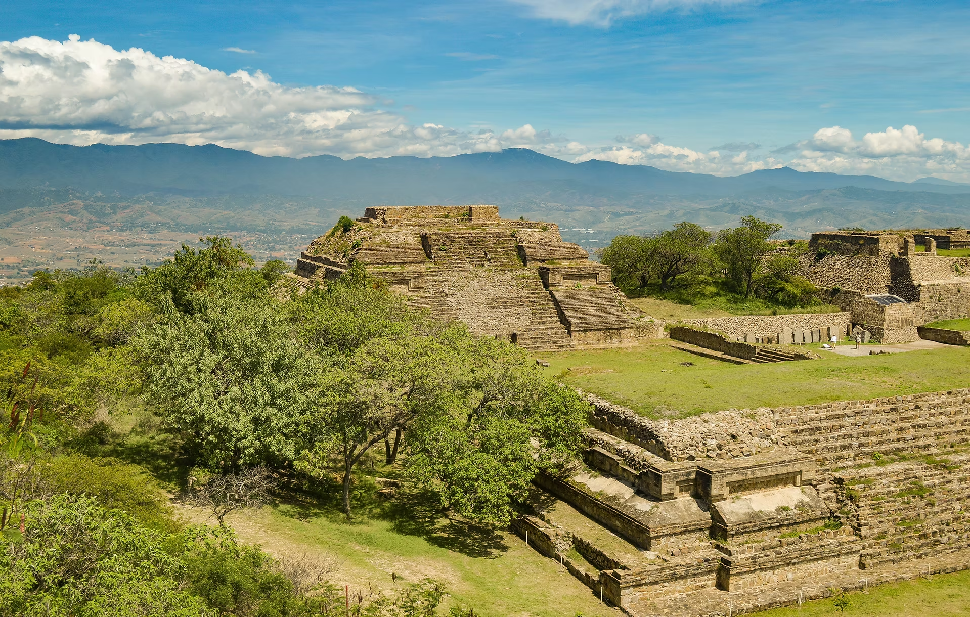 Ruines de pyramides anciennes au milieu d'une végétation verte avec des montagnes et un ciel bleu avec des nuages en arrière-plan.