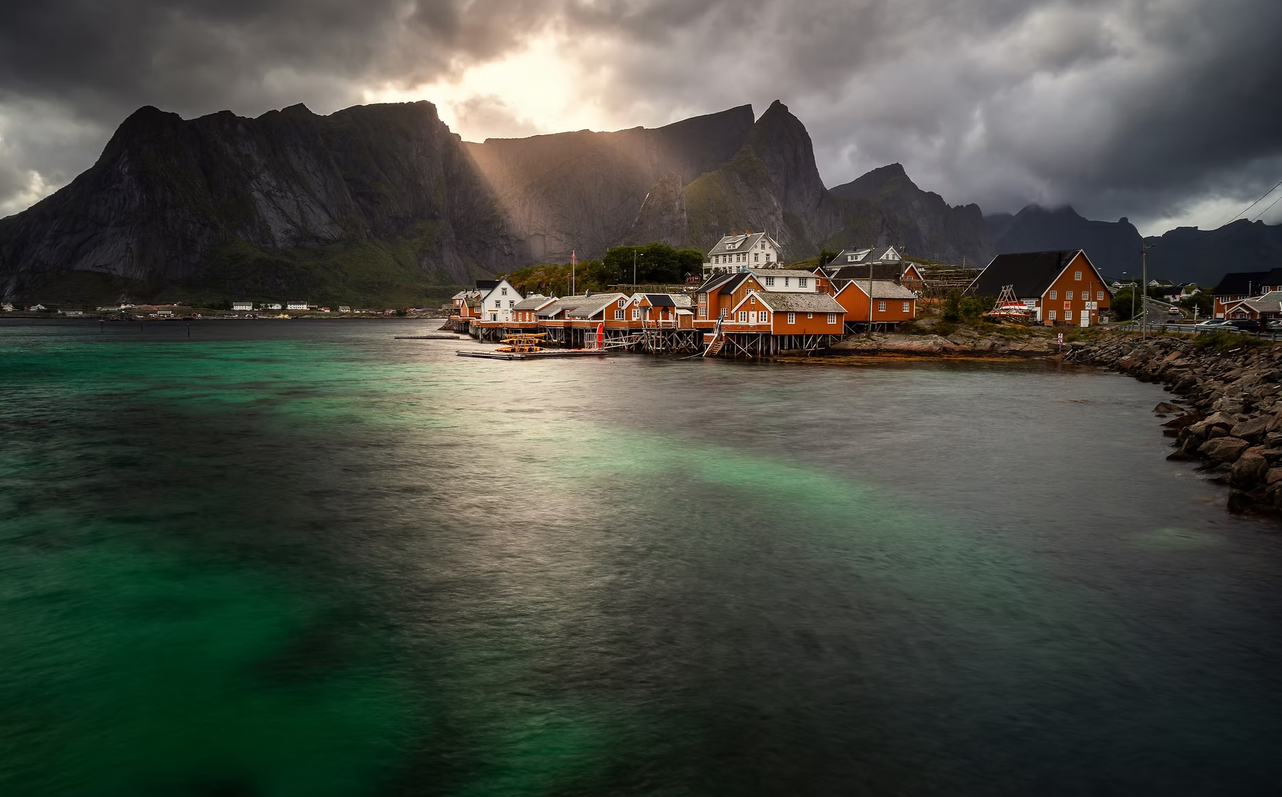 Villages de pêcheurs colorés en bois sur l'eau calme, grandes montagnes rocheuses en arrière-plan sous un ciel nuageux avec des rayons de soleil filtrant à travers les nuages.