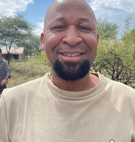 Un homme souriant avec un environnement rural en arrière-plan, arbres et ciel nuageux.