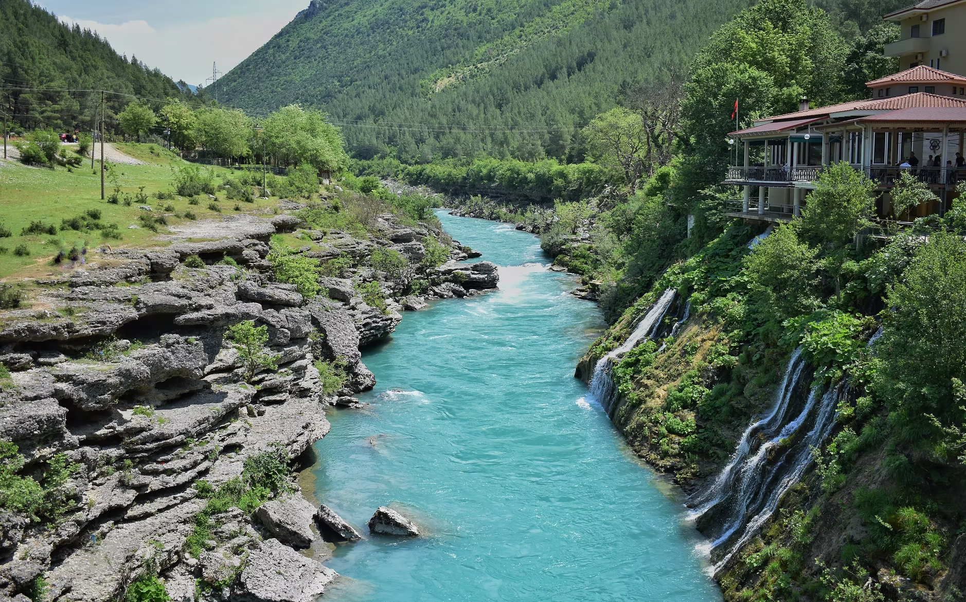 Une rivière bleue coule entre des rochers et des arbres verts, avec une maison en balcon sur la droite, entourée de montagnes et de forêt dense.