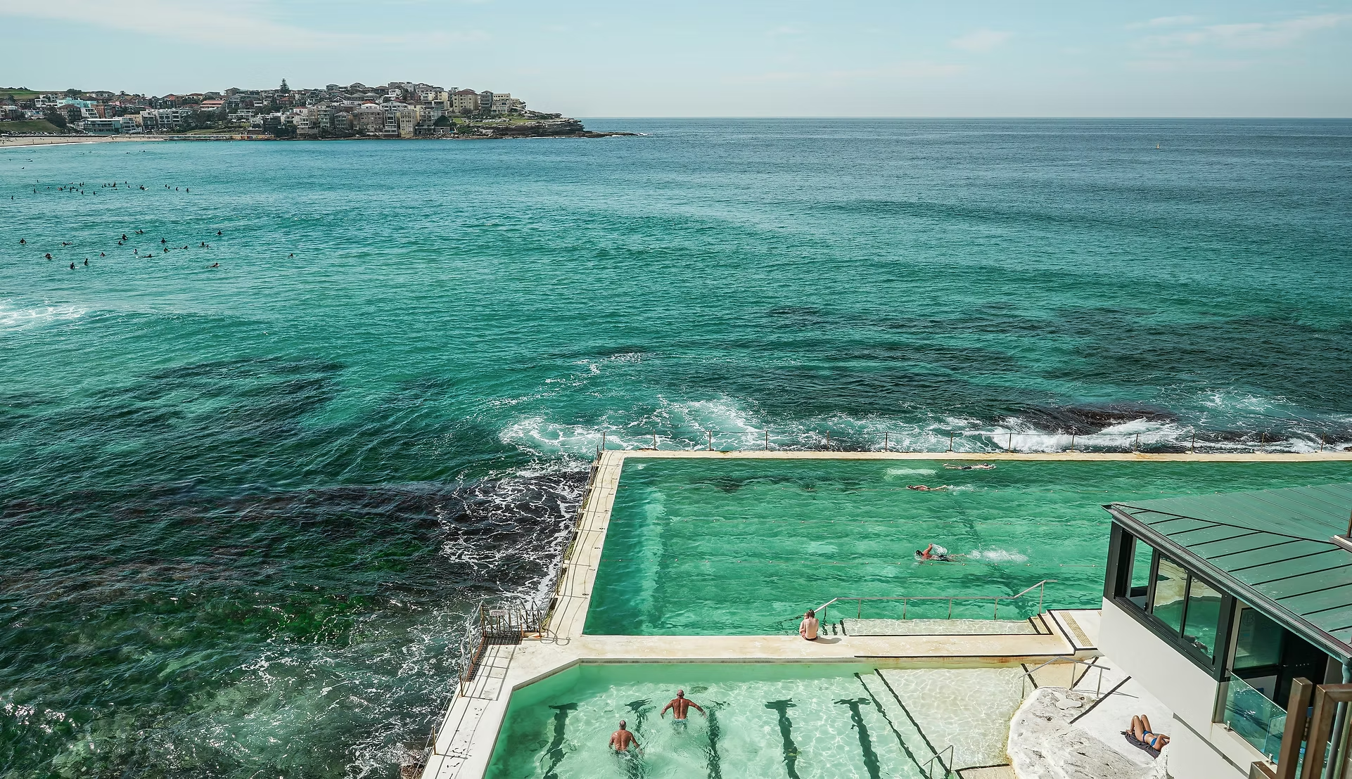 Piscine à débordement située au bord de la mer avec des personnes nageant à l'intérieur, vue sur l'océan et la côte en arrière-plan.