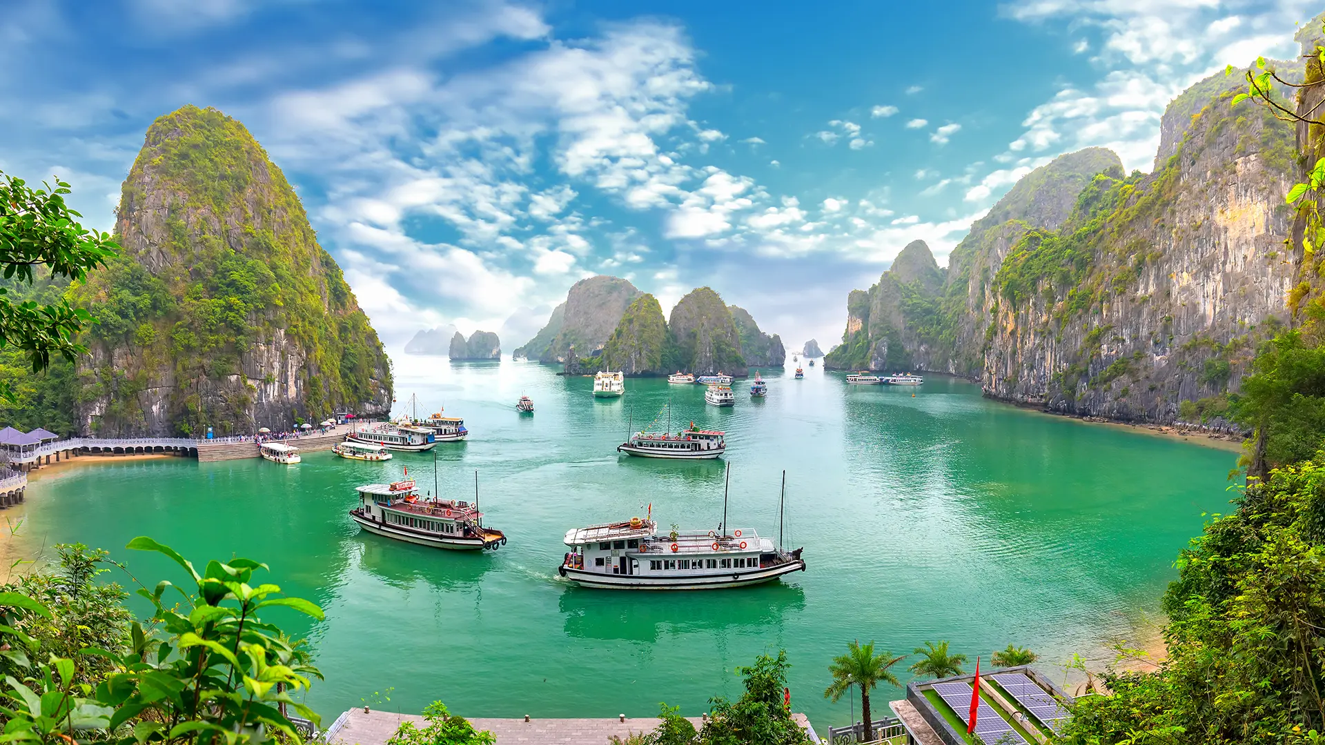 Une baie avec plusieurs montagnes karstiques recouvertes de végétation, un ciel bleu avec quelques nuages, et plusieurs bateaux de tourisme flottant sur une eau turquoise. Il y a aussi une promenade en bois le long du rivage et des parasols sous un ciel ensoleillé.