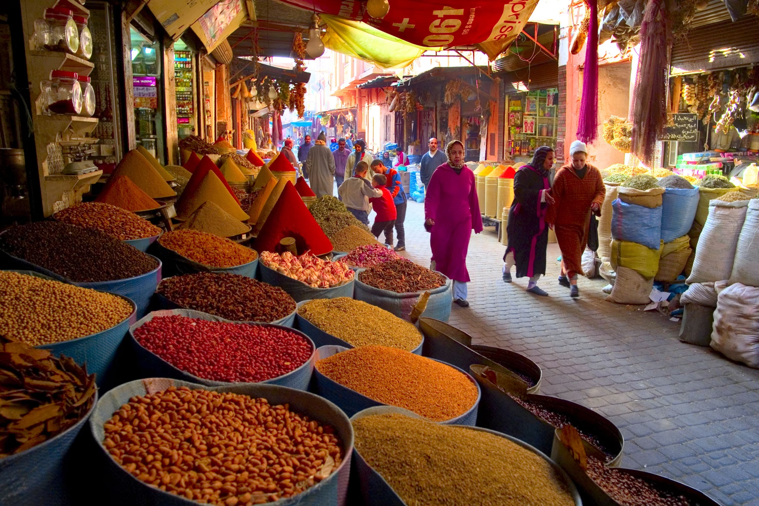 Un marché coloré avec des sacs de différentes épices et herbes, des vendeurs et des acheteurs marocains dans une ruelle étroite, sous des tentes colorées.