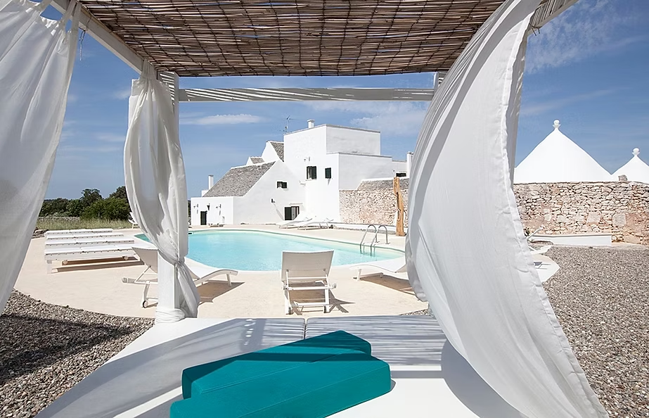 Vue d'une piscine entourée de bâtiments blancs dans un complexe hôtelier, vue depuis une cabane en bambou avec rideaux blancs.