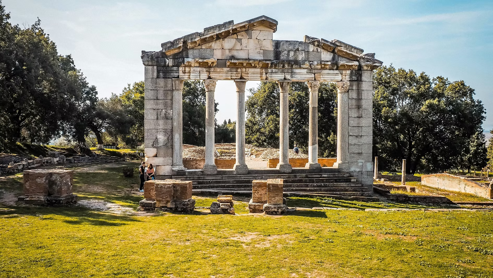 Ruines antiques avec colonnes dans un parc entouré d'arbres, trois personnes discutant devant la structure, ciel clair et soleil