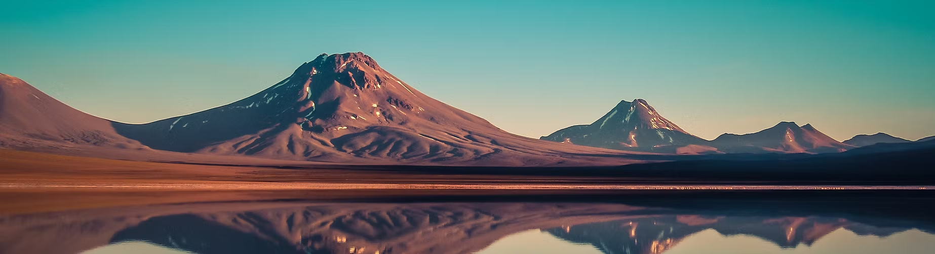 Paysage de montagnes volcaniques avec reflets dans un lac calme au premier plan. Le ciel est clair et en dégradé de bleu.
