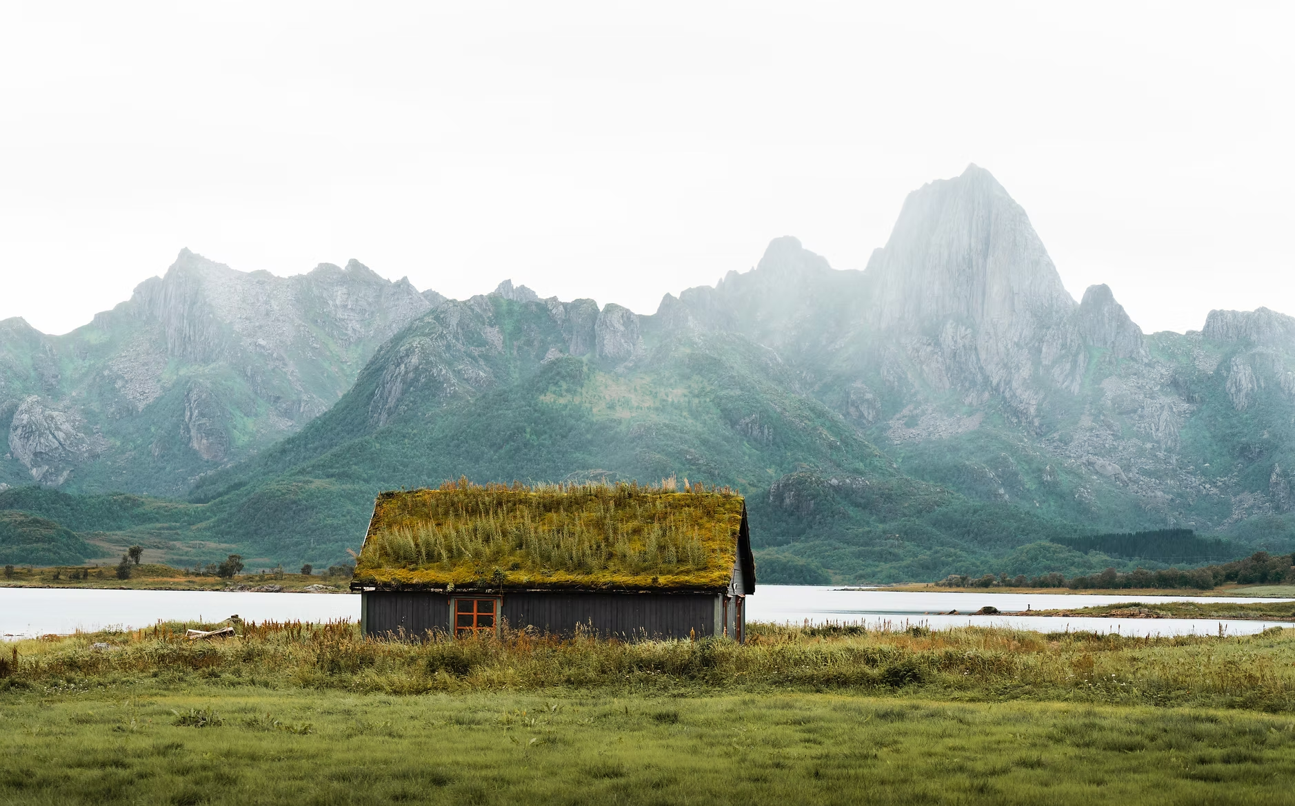 Une cabane en bois avec un toit recouvert de mousse, située au bord d’un lac, avec des montagnes verdoyantes en arrière-plan sous un ciel nuageux.