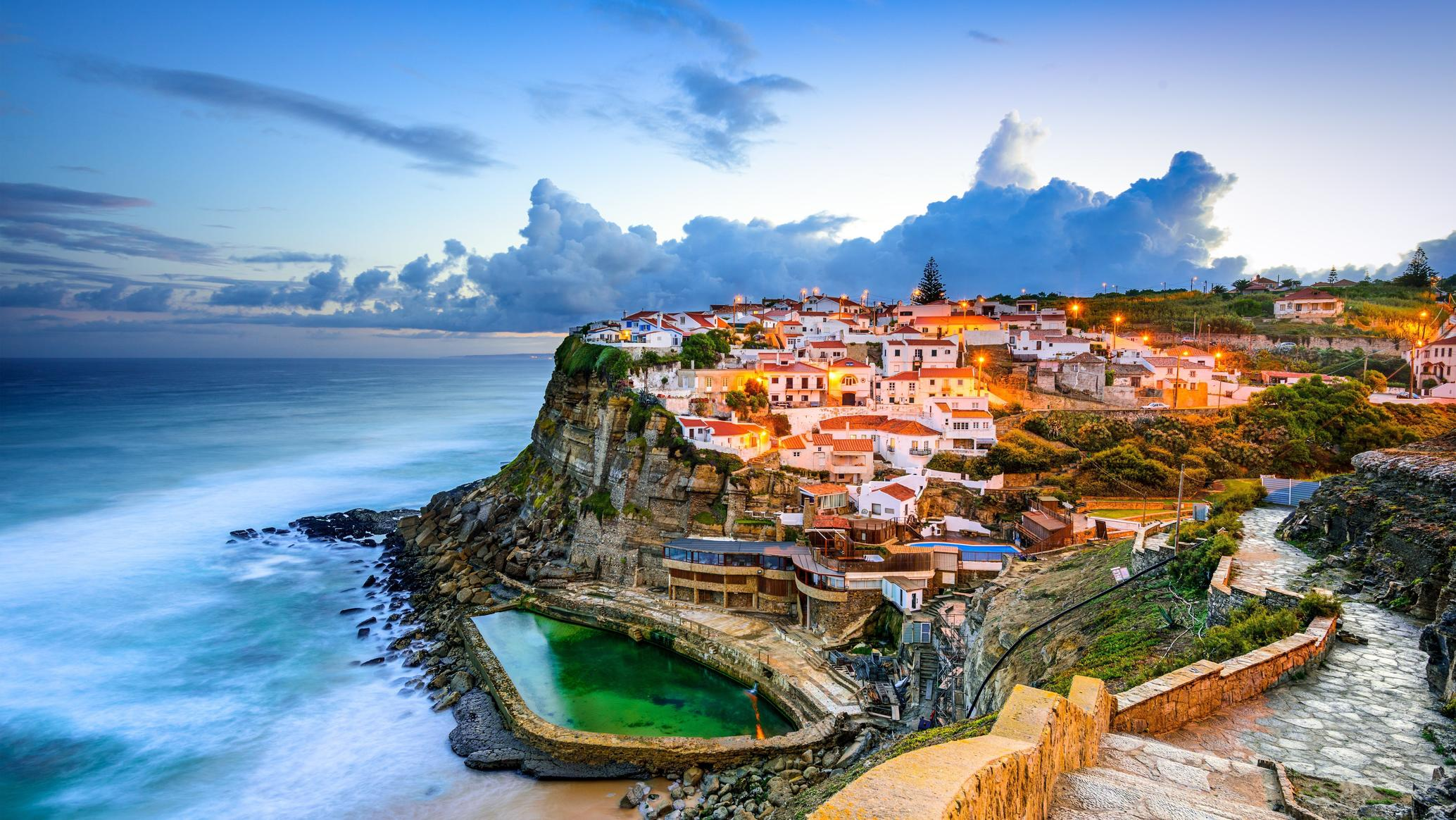 Vue d'une ville côtière avec maisons blanches perchées sur une falaise, au coucher du soleil, avec la mer à gauche et un ciel orageux en arrière-plan.