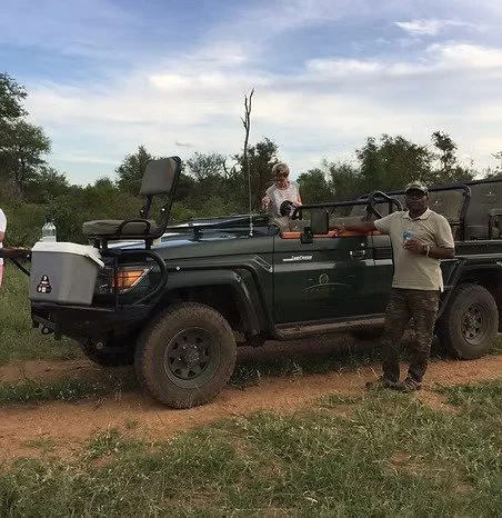 Deux personnes se tiennent près d'un véhicule tout-terrain dans un environnement naturel avec des arbres et un ciel partiellement nuageux.