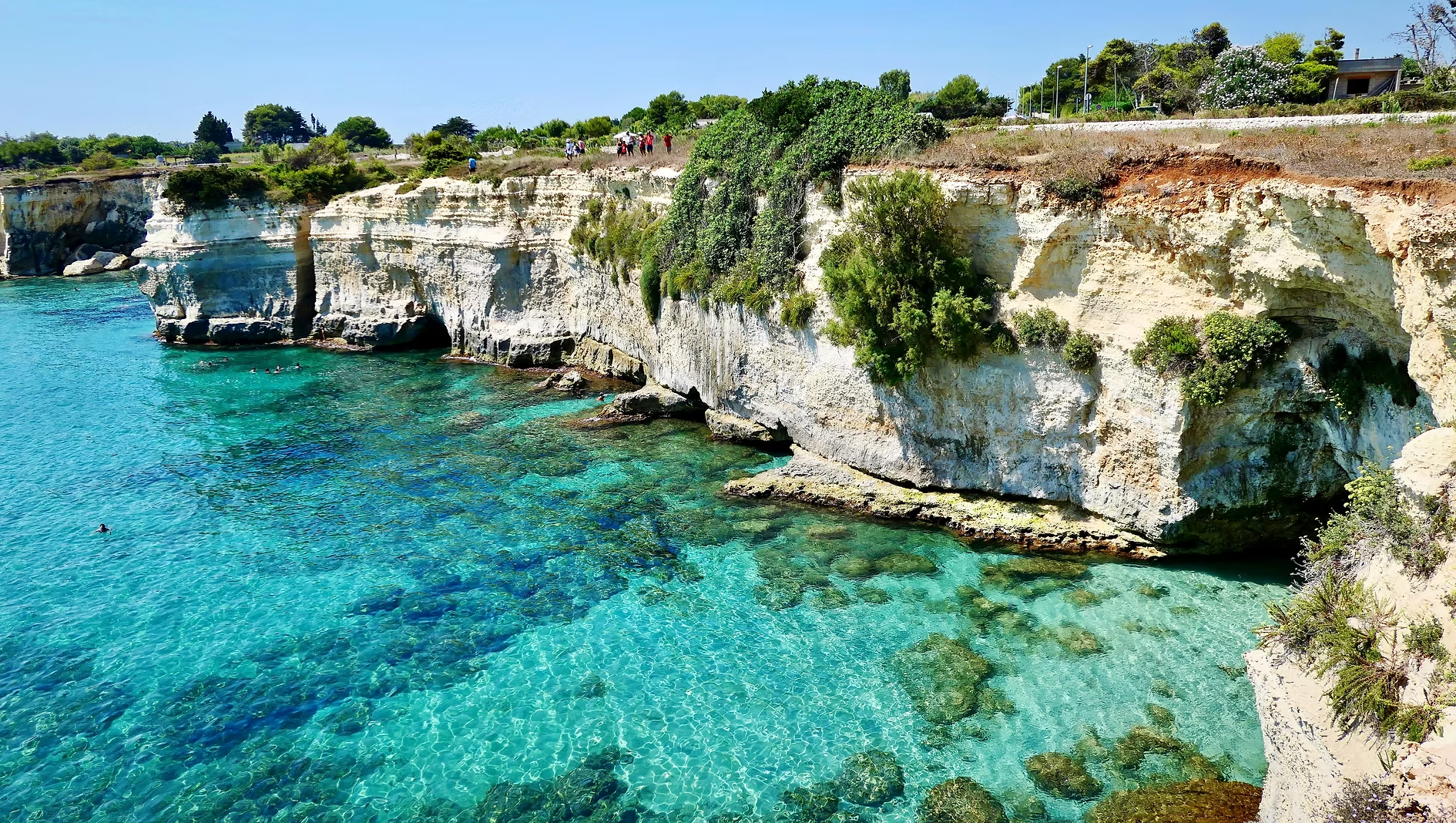 Criques rocheuses avec eau turquoise claire, vue sur la mer et la côte avec des plantes et des arbres, quelques personnes nageant et marchant sur la falaise.