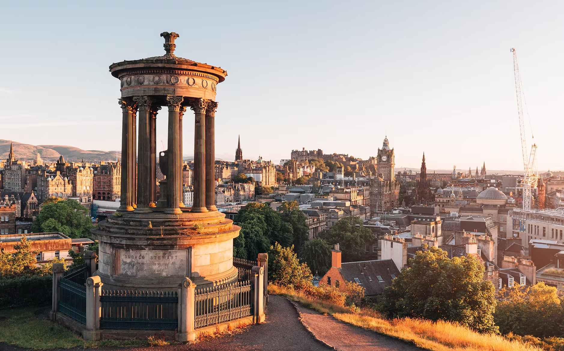 Paysage urbain de Édimbourg au coucher du soleil, avec une vue sur la ville et le château d'Édimbourg à l'arrière-plan. En premier plan, un monument en pierre avec des colonnes et une plaque à son socle.