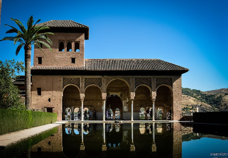Un bâtiment historique avec un patio ensoleillé, arches et miroir d'eau, entouré de végétation et de collines en arrière-plan.