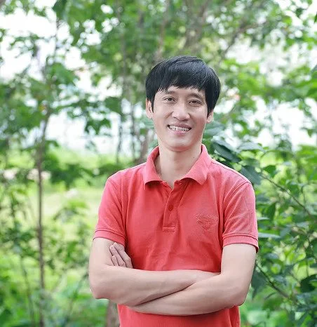 Un homme souriant portant un polo rouge, debout dans un jardin vert avec des arbres en arrière-plan.