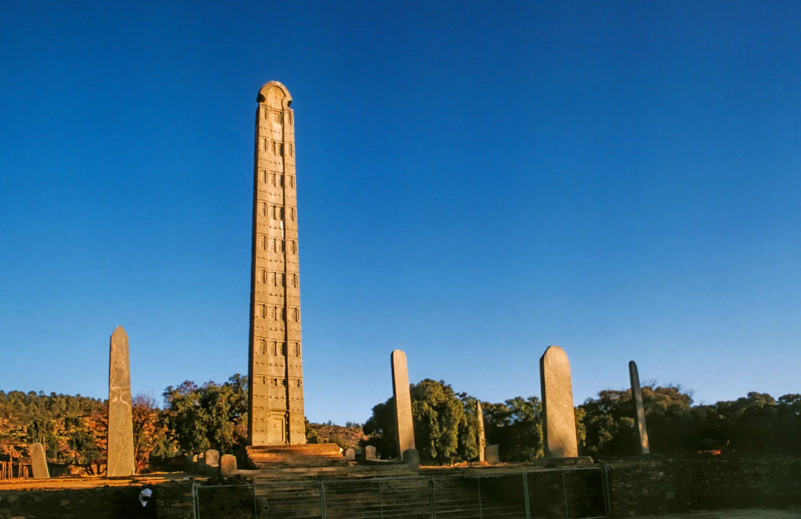Un obélisque ancien entouré de plusieurs pierres et d'arbres, sous un ciel clair et ensoleillé.