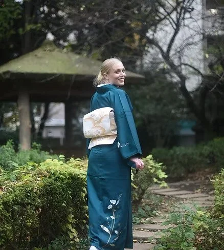 Femme portant un kimono bleu avec un obi beige, se tenant dans un jardin avec des arbres et un chemin en pierre.