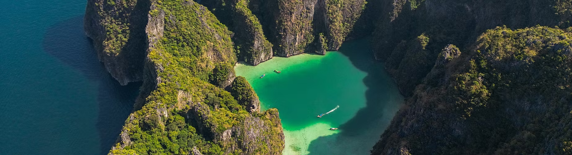 Vue aérienne d'une baie entourée de falaises vertes avec de l'eau turquoise et des bateaux naviguant.