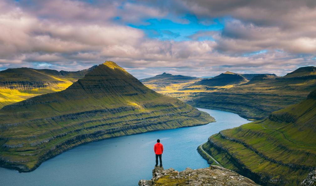 Une personne en manteau rouge regarde un canyon avec une rivière serpentante, entouré de montagnes verdoyantes sous un ciel nuageux.
