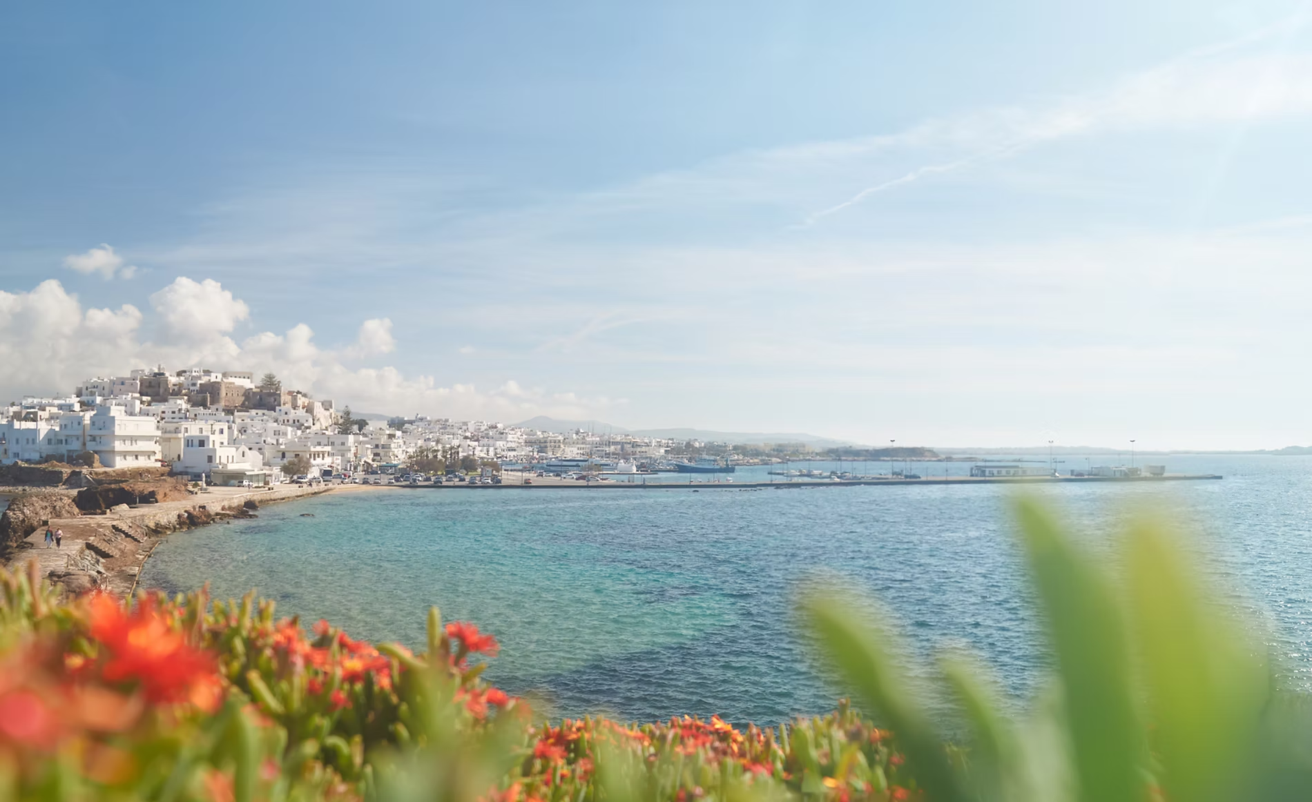 Vue panoramique d'une ville côtière avec bâtiments blancs, un port avec bateaux, la mer calme et un ciel en partie nuageux.