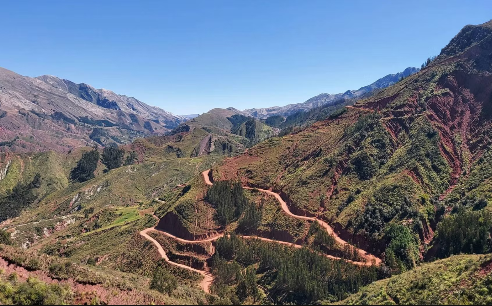 Paysage de montagnes avec un sentier sinueux rougeâtre et des collines verdoyantes sous un ciel clair et bleu.