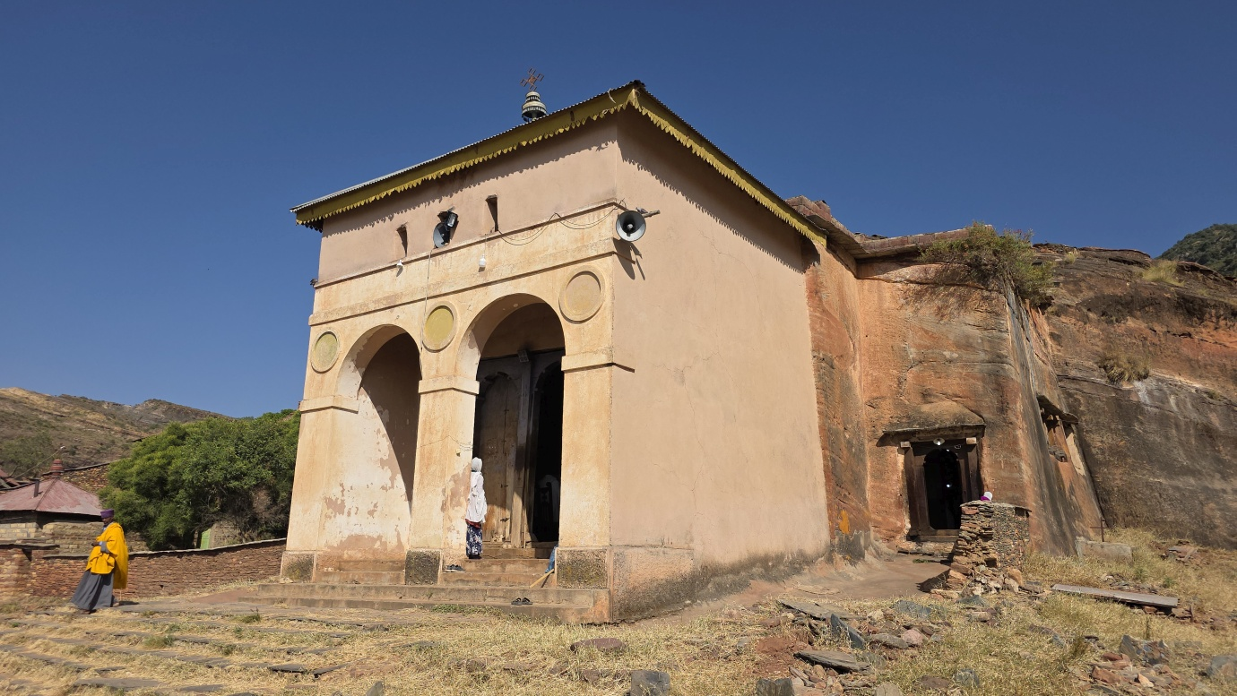 Une vieille église en pierre, située dans un paysage de montagnes arides, avec deux personnes devant l'entrée.
