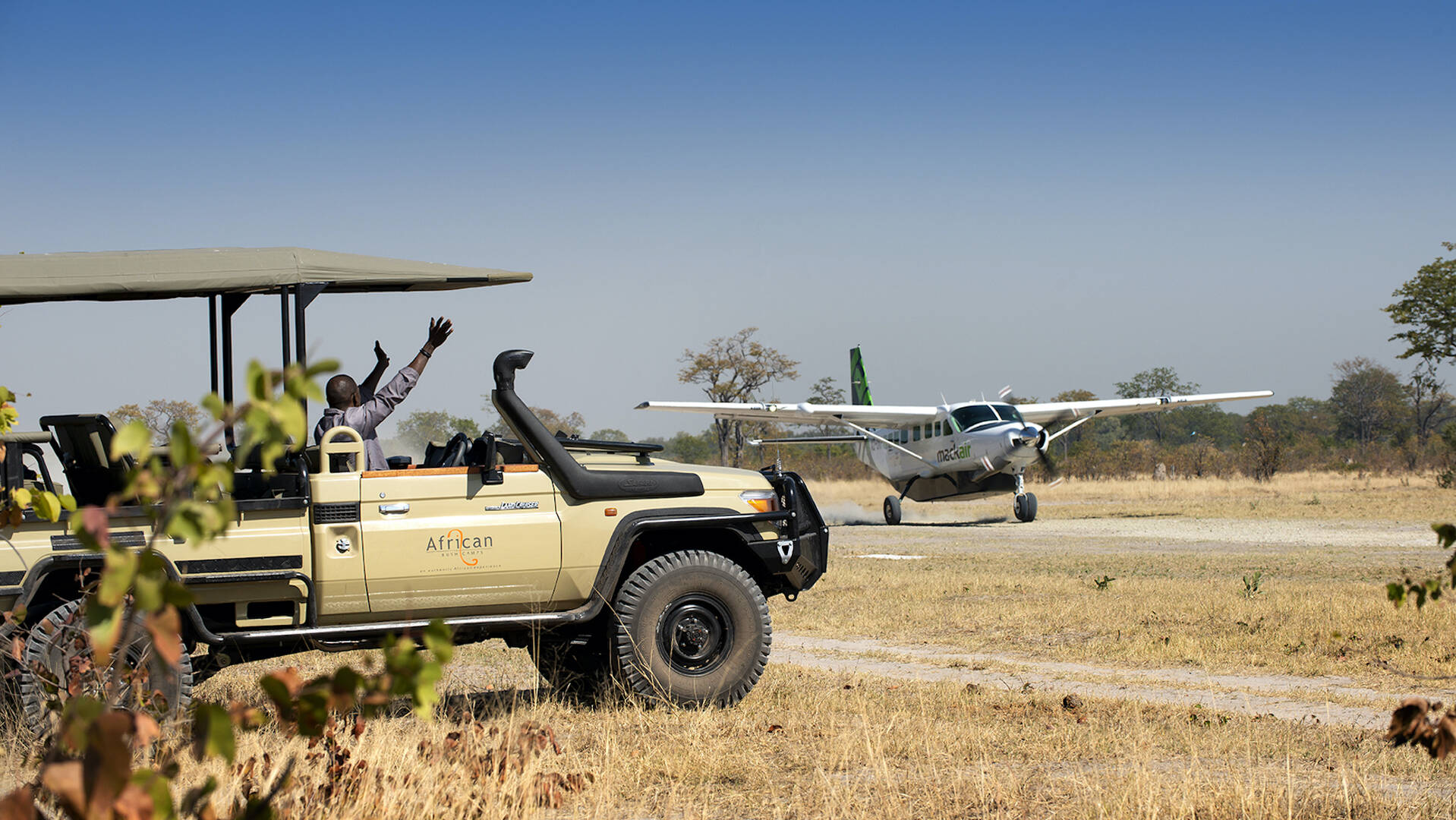 Une personne dans un véhicule de safari salue vers un petit avion qui atterrit sur une piste en zone de savane africaine.