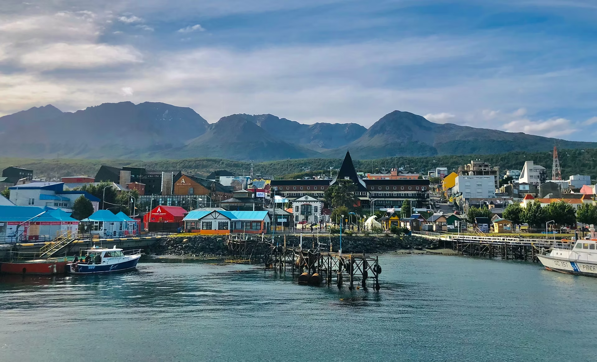 Un port avec des bateaux, un village coloré avec des bâtiments variés et des montagnes au loin sous un ciel partiellement nuageux.
