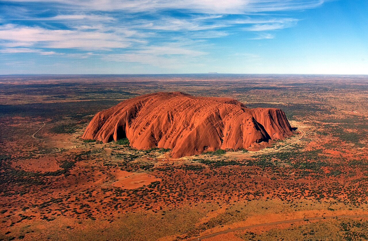 Une vue aérienne de Uluru, un monolithe rouge situé dans le centre de l'Australie, avec un ciel partiellement nuageux.