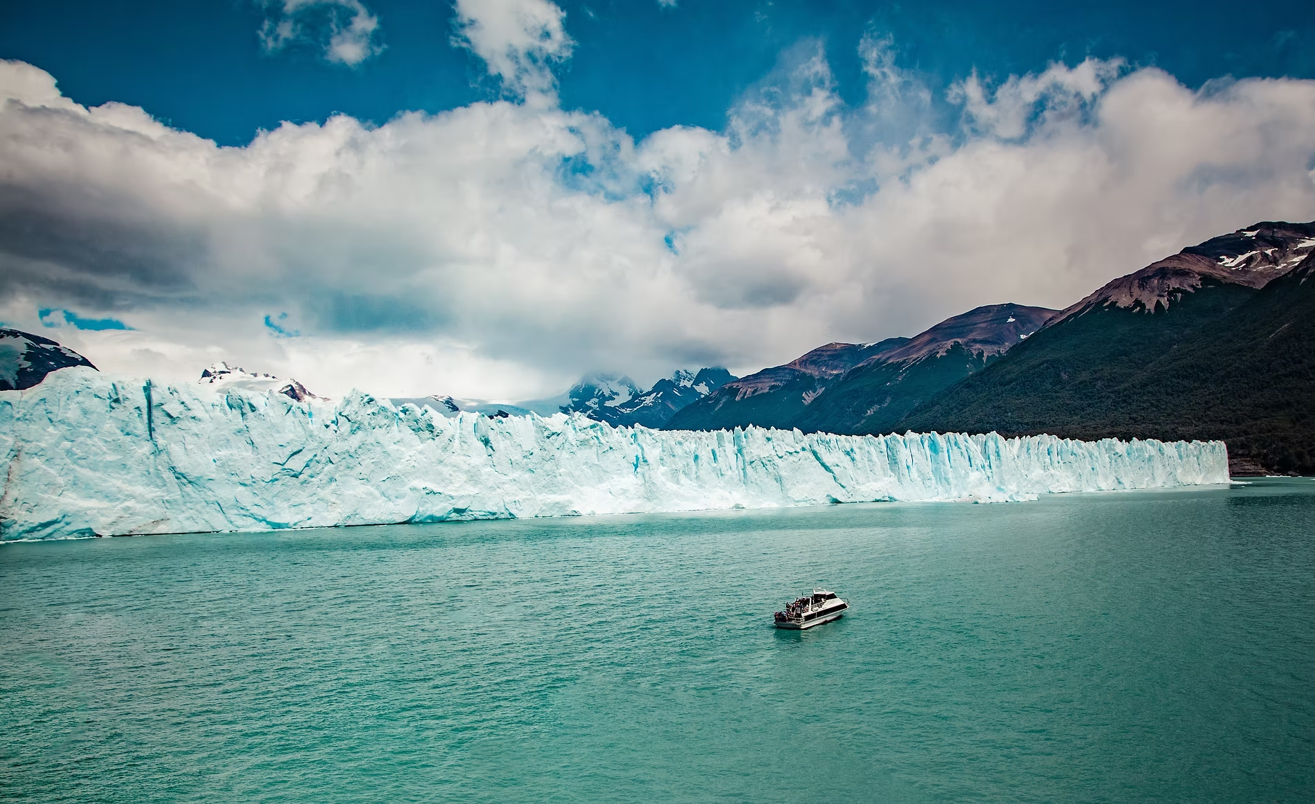 Un glacier bleuté s'étend dans un lac calme avec des montagnes en arrière-plan et des nuages dans le ciel.