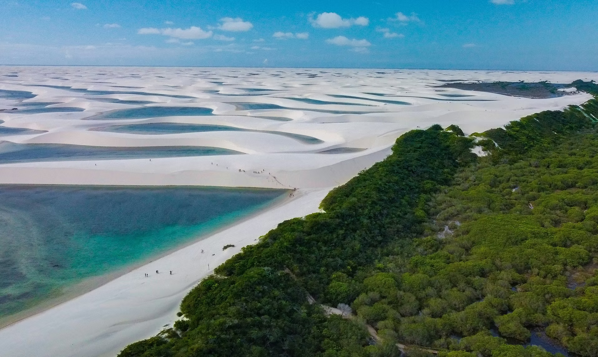 Une vue aérienne de dunes de sable blanc avec des parties d'eau turquoise, entourées de végétation verte, sous un ciel bleu avec quelques nuages.