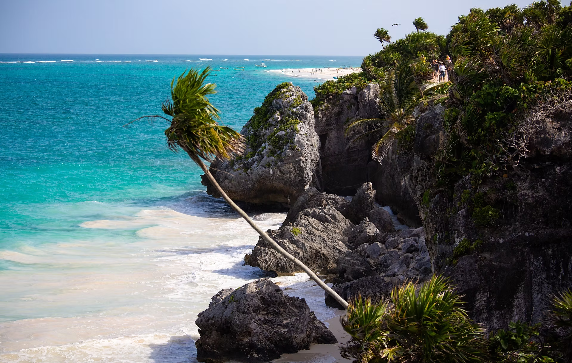Une côte rocheuse tropicale bordée par la mer turquoise. Des végétations verdoyantes, dont un palmier penché, poussent sur les rochers. Au loin, une plage de sable blanc avec des personnes se promenant et quelques bateaux.