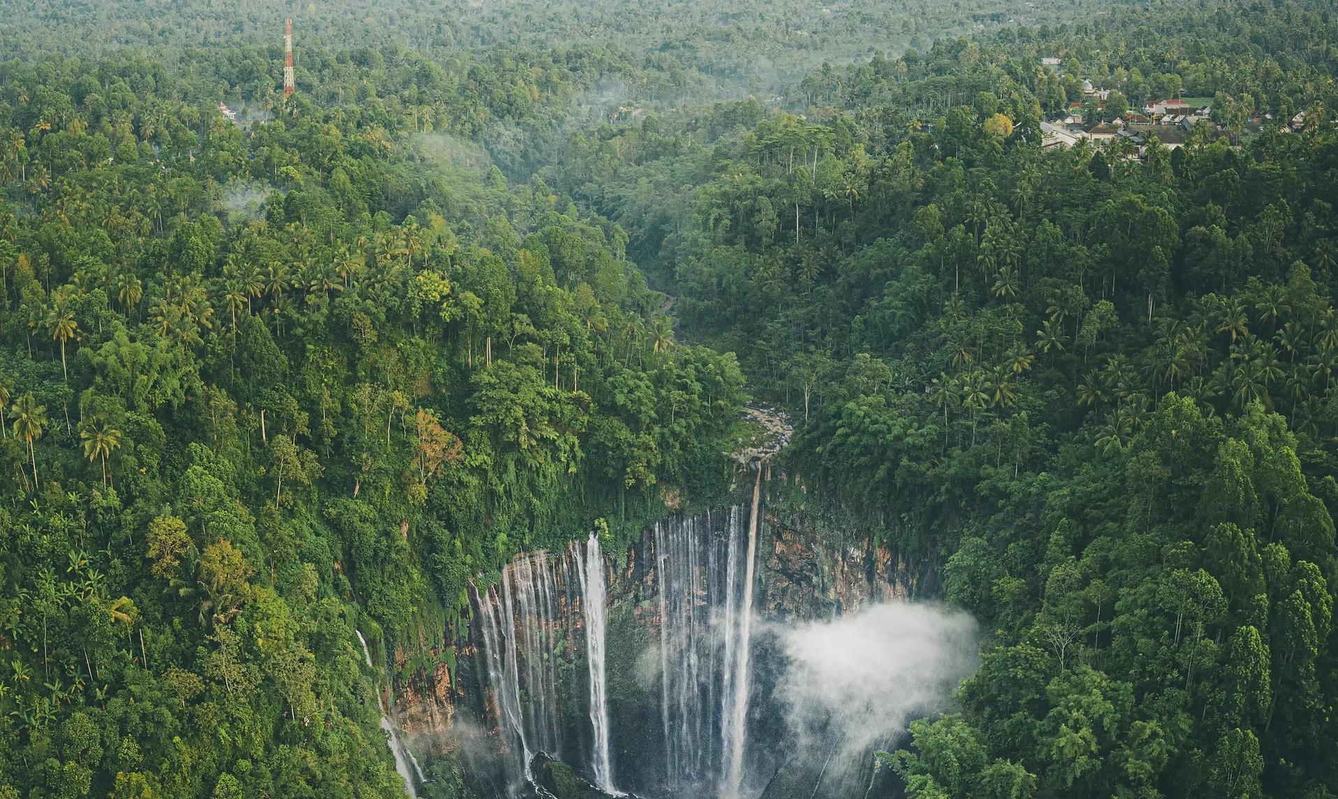 Une cascade entourée d'une dense forêt tropicale verte, avec quelques bâtiments visibles au loin et une tour en haut à gauche.