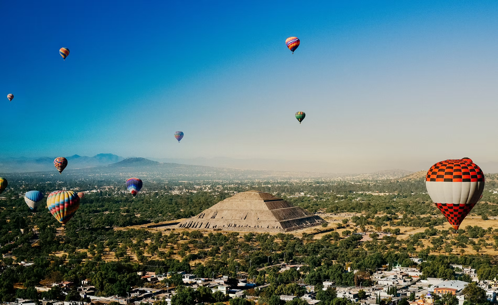 De nombreux ballons à air chaud colorés flottent dans le ciel clair au-dessus d'une ville et des pyramides."
