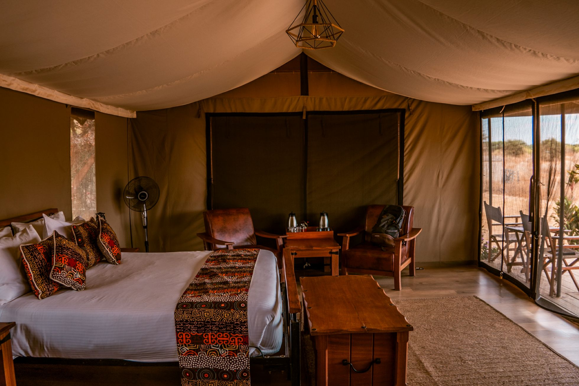 Intérieur d'une tente de camping deluxe avec un lit fait, un fauteuil en bois, une petite table, une lampe suspendue en métal et des fenêtres avec vue sur un paysage aride.