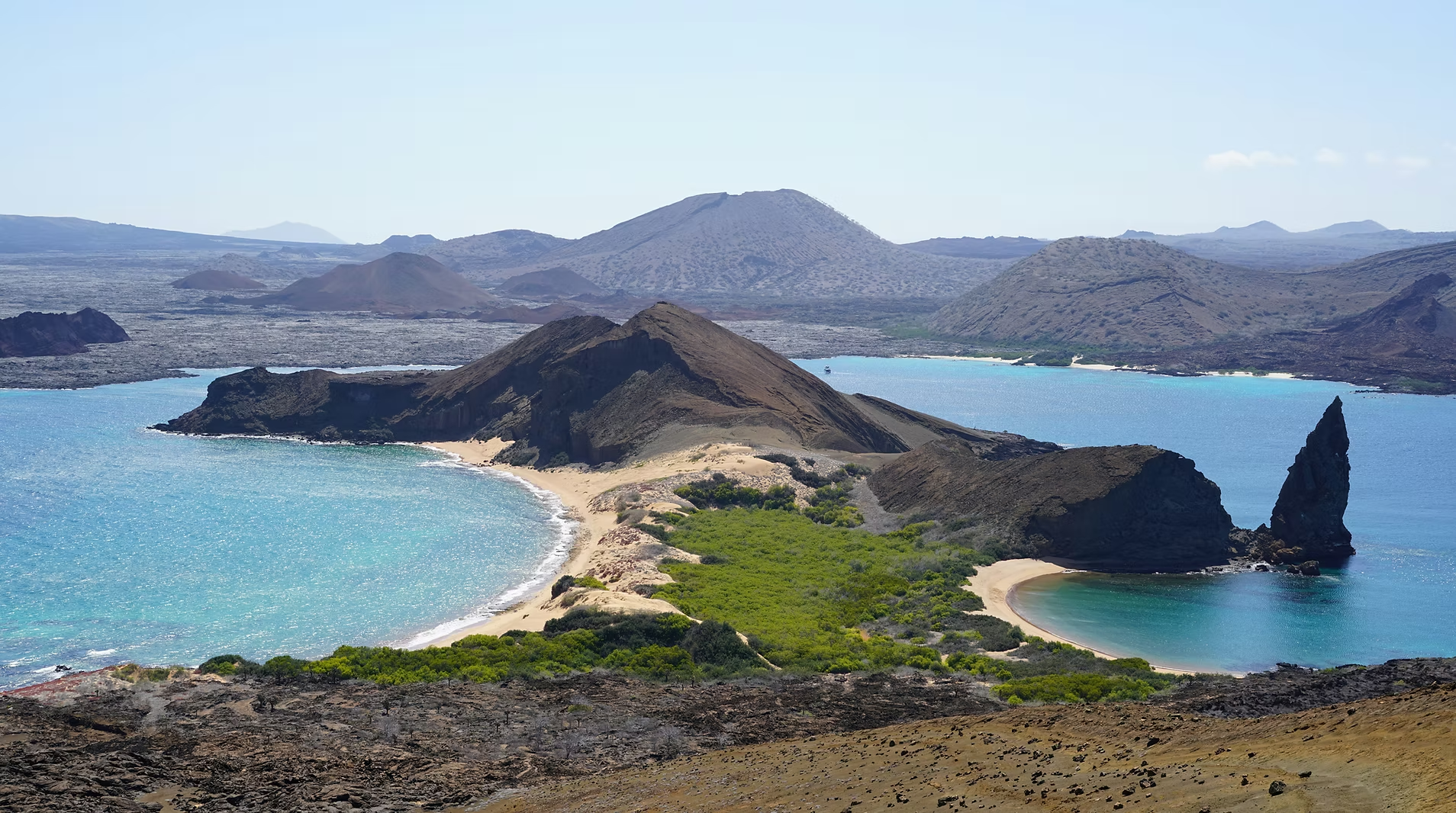 Paysage côtier avec plage, eau turquoise, formations volcaniques noires, montagnes en arrière-plan et végétation verte.