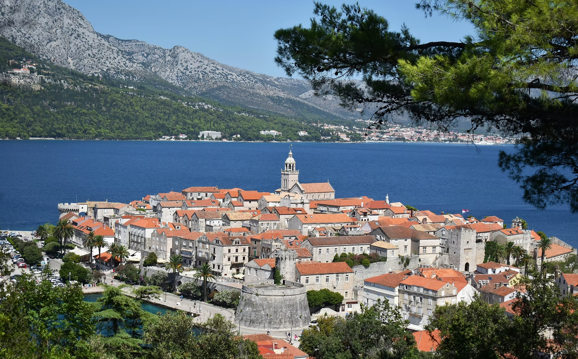 Une vue d'une ville côtière méditerranéenne avec des bâtiments en pierre aux toits en tuiles rouges, une tour d'église, et un lac ou mer bleu entouré de montagnes, avec des arbres verts encadrant l'image.
