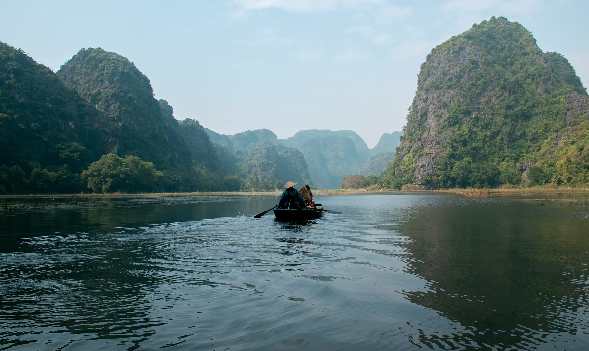 Une barque avec deux personnes navigue sur une rivière entourée de montagnes verdoyantes dans un paysage naturel paisible.