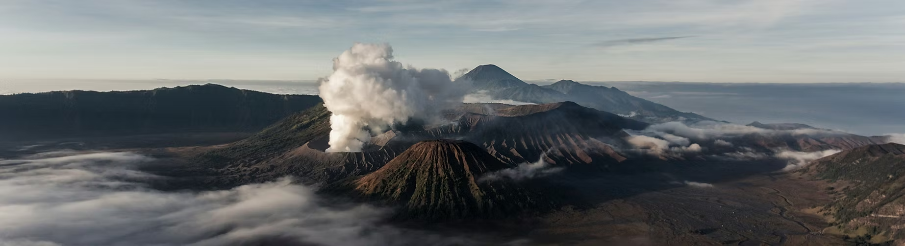 Vue aérienne d'un volcan en activité avec de la fumée s'échappant, entouré de montagnes et de nuages.