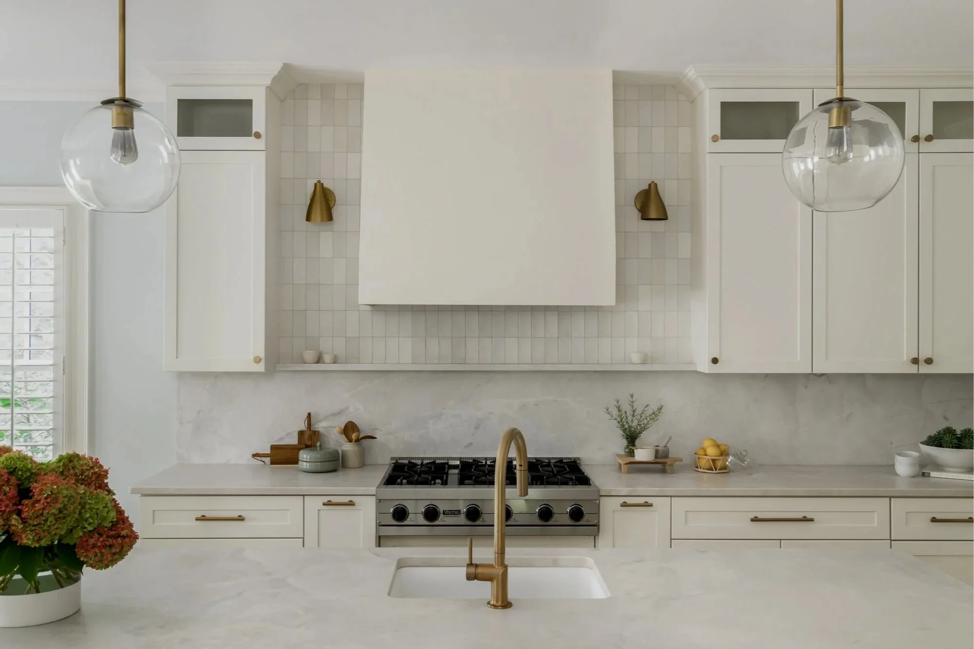 A modern white kitchen with a marble countertop, gold hardware, white cabinetry, and gold pendant lights. There is a flower arrangement on the counter, and a stove beneath a white tiled backsplash.