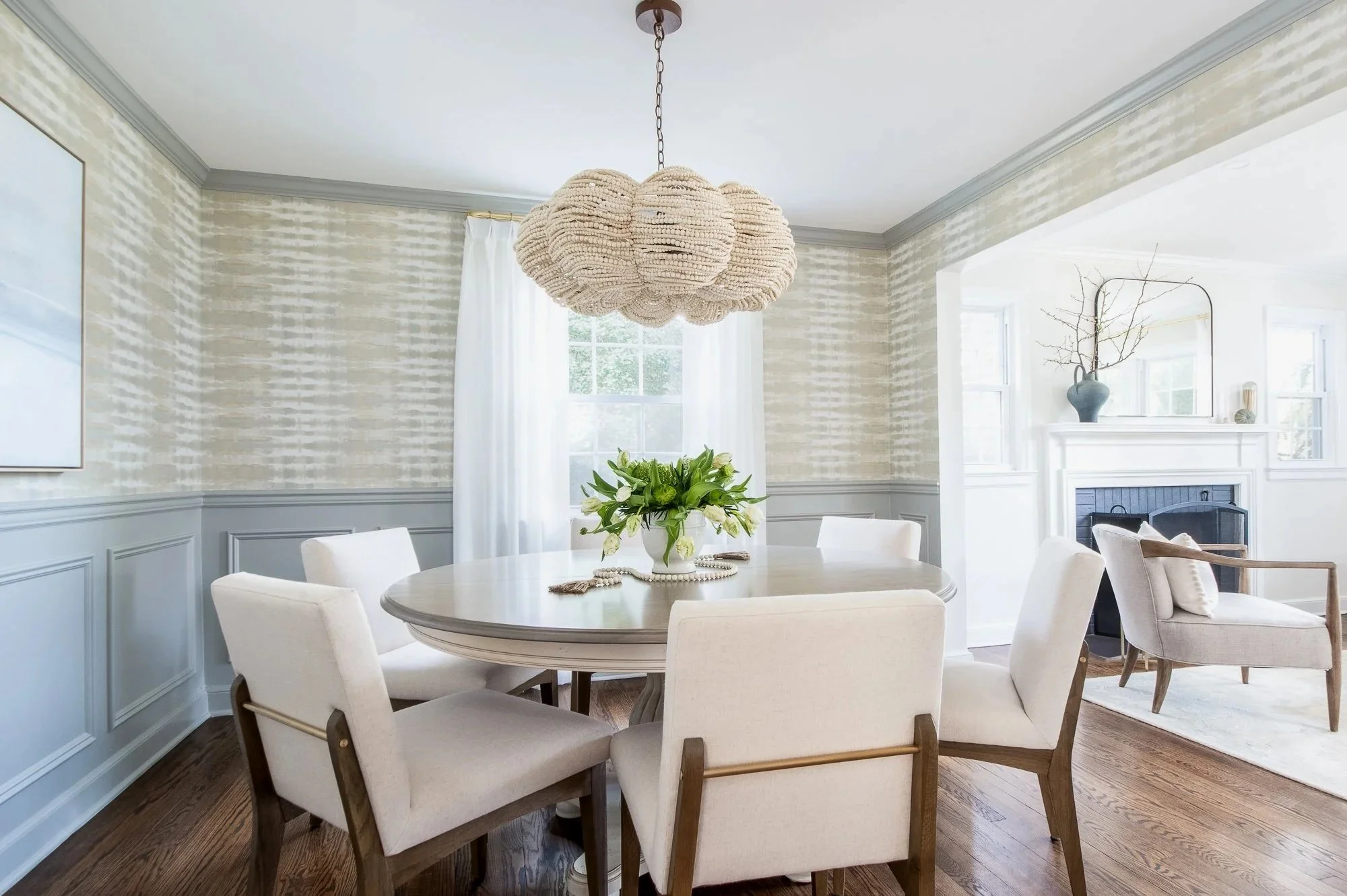 Bright dining room with a round table surrounded by six white upholstered chairs. A floral centerpiece with white flowers sits on the table. Pendant chandelier made of shells hangs from the ceiling. Light-colored walls with wainscoting and large wind