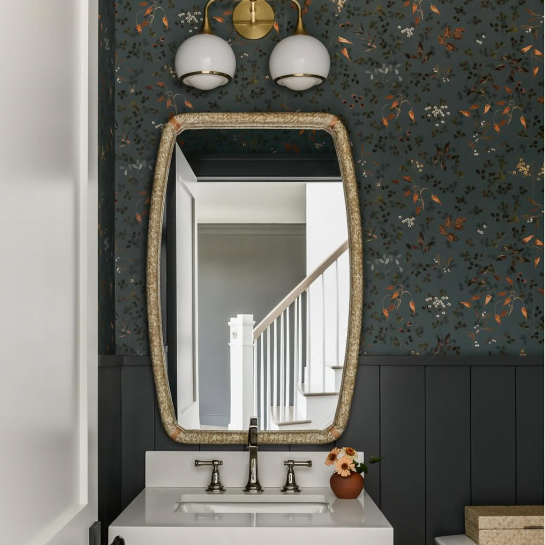 Decorative bathroom mirror above a white sink, dark floral wallpaper, wall sconces, and a small vase with flowers on the sink