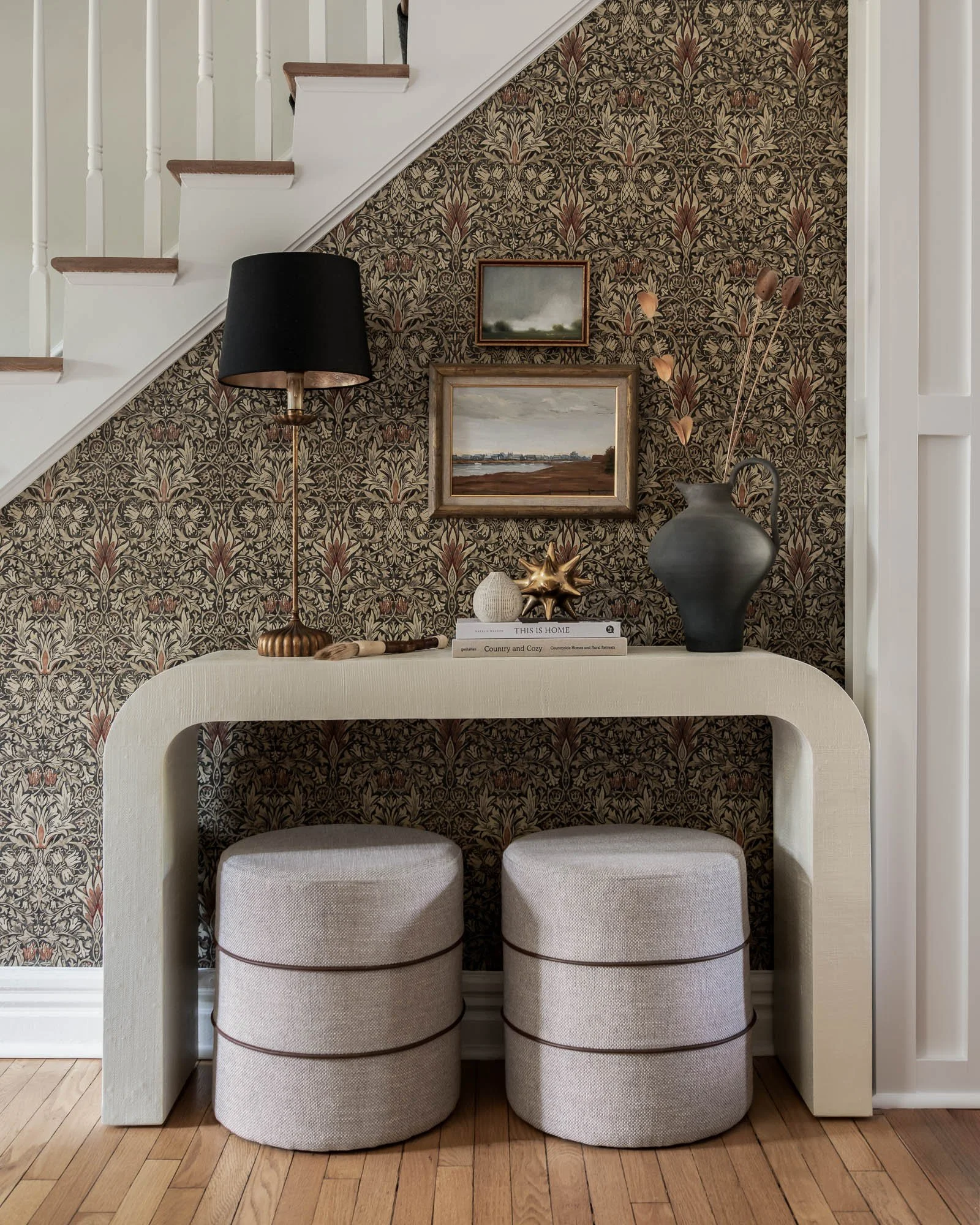 Interior of a cozy living space featuring a decorative wallpapered wall with two framed landscape paintings. A black table lamp with a gold base is next to a large black vase with dried branches. Two neutral-colored upholstered ottomans are positioned in front of the wall, and a stack of books with decorative objects is on a small table beneath the wall.