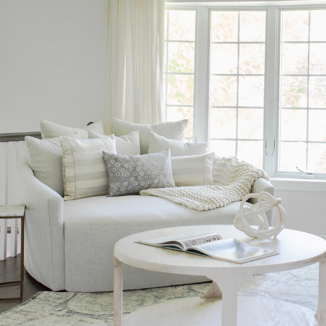 Bright living room with a white sofa topped with multiple white and gray pillows, a cream throw blanket, next to a large window with white curtains. A round white coffee table holds an open book, a magazine, and a decorative white sculpture. Light filters through with soft natural illumination.