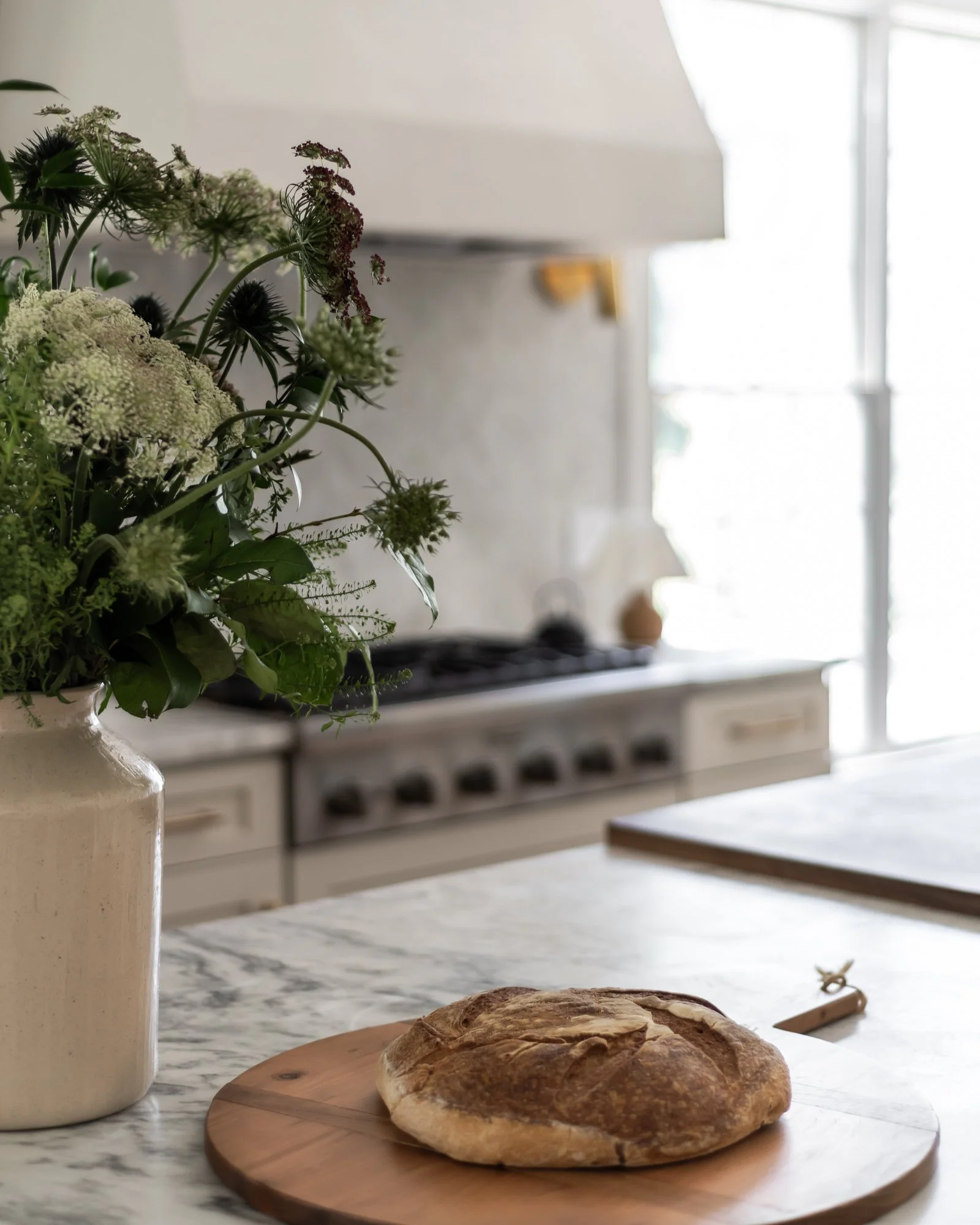 Fresh loaf of bread on a wooden cutting board on a marble kitchen counter, with a knife nearby, in a bright kitchen with natural light coming through windows.