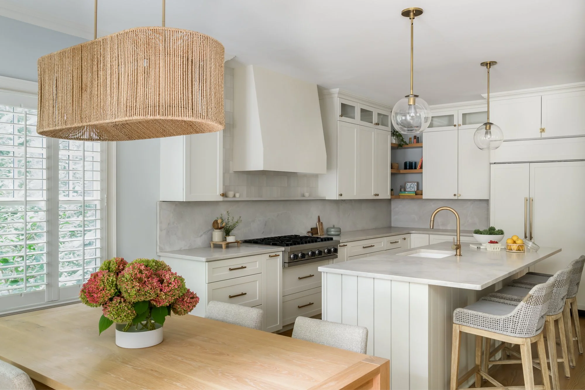 A modern kitchen with white cabinets, a marble backsplash, and a kitchen island. There are two glass pendant lights hanging from the ceiling, and a large window with white shutters. A dining table with a potted pink hydrangea plant is in the foregrou