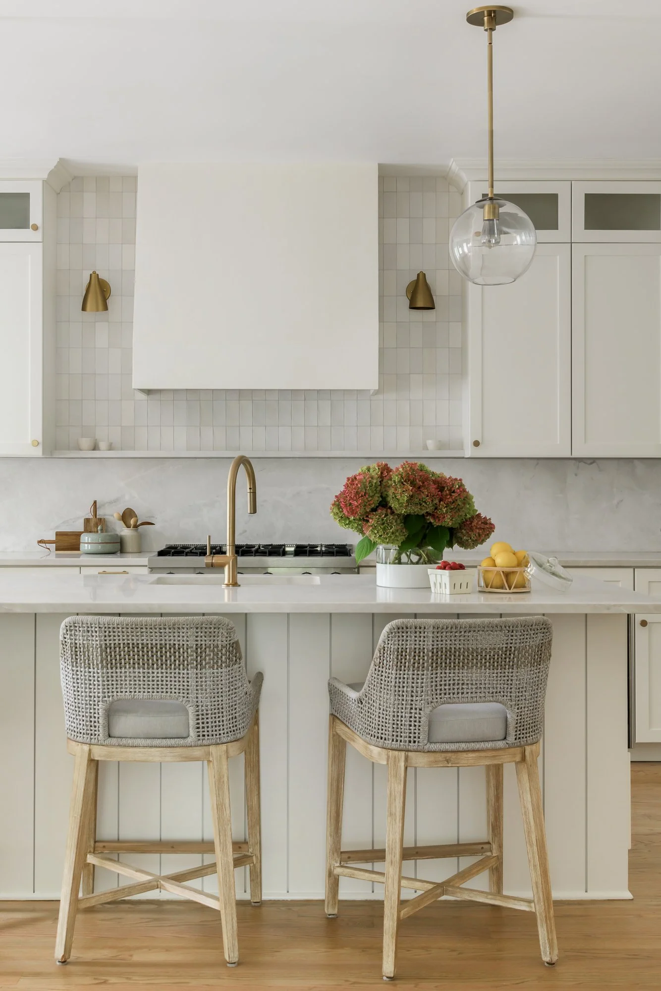 A kitchen with a white island counter, two woven back chairs, a flower centerpiece, and gold accents including a faucet, wall sconces, and a hanging light fixture.